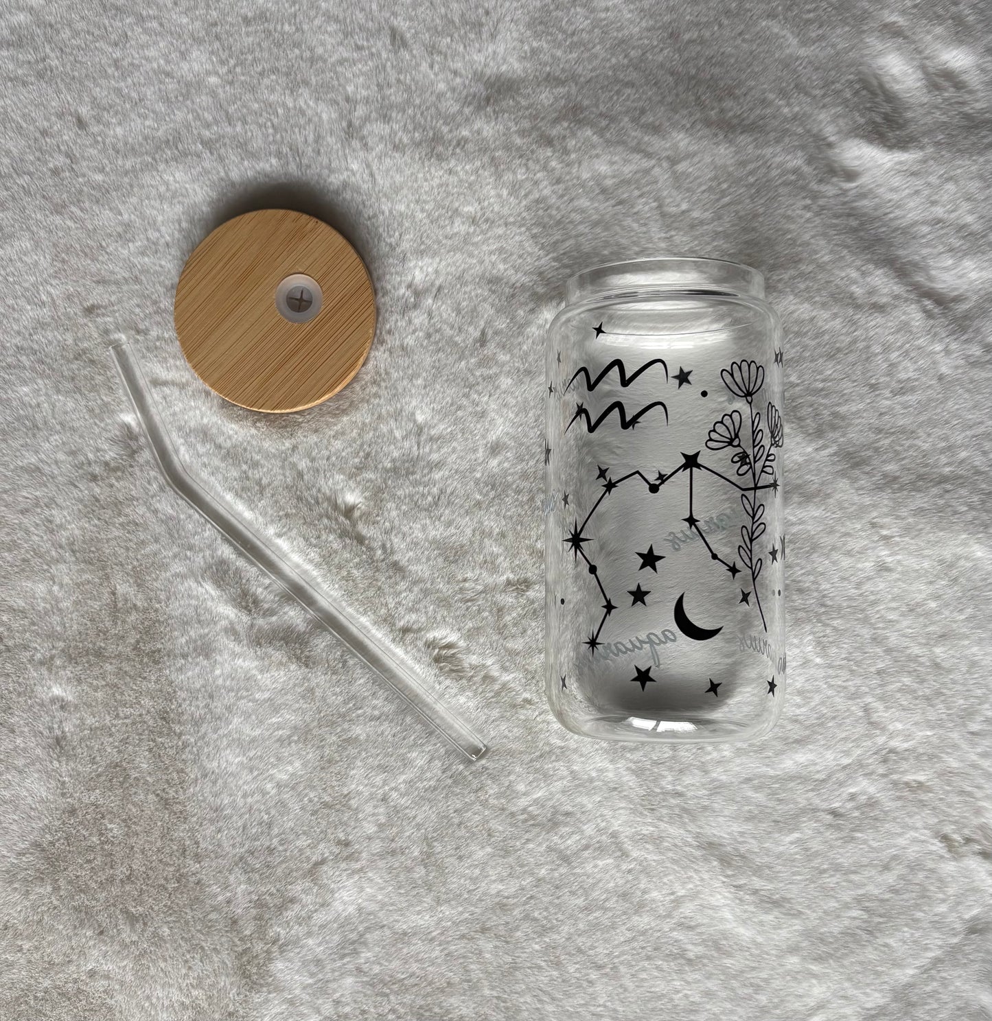 Glass can cup with glass straw and bamboo lid. Design on the glass cup is the Aquarius zodiac symbol, star constellation, flower and the text Aquarius on the backside