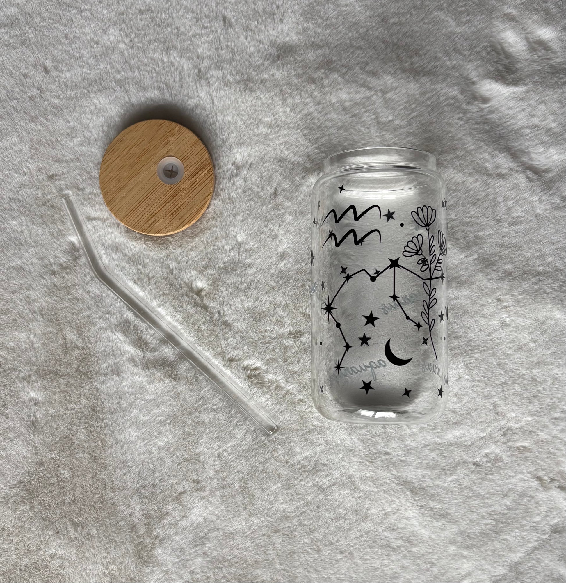 Glass can cup with glass straw and bamboo lid. Design on the glass cup is the Aquarius zodiac symbol, star constellation, flower and the text Aquarius on the backside